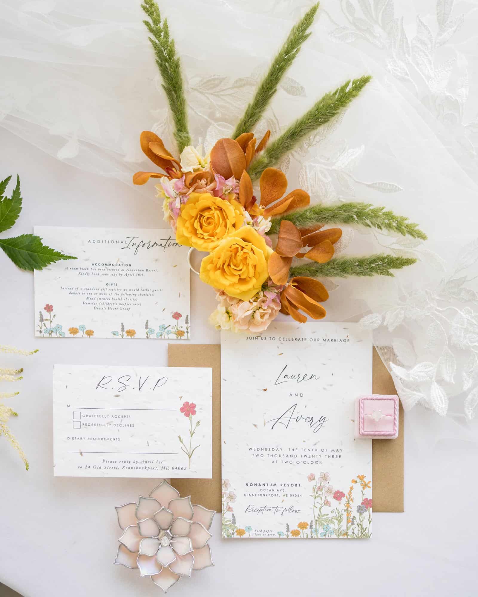 A wedding invitation suite featuring RSVP and details cards, next to a bridal bouquet with yellow and orange flowers, and a ring in an open pink box.