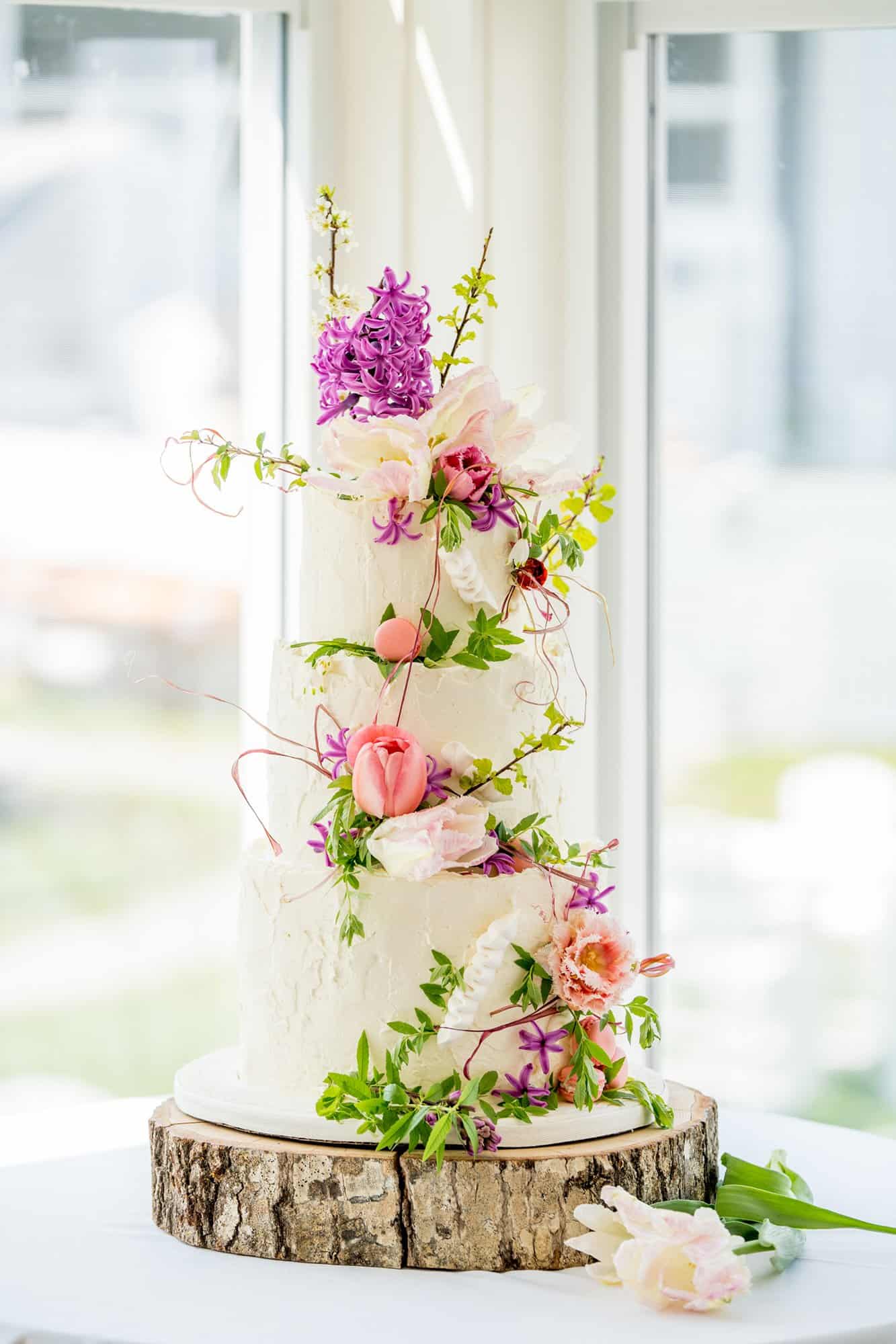 A three-tier white cake adorned with pink and purple flowers and green foliage, displayed on a round wooden base against a background of windows.