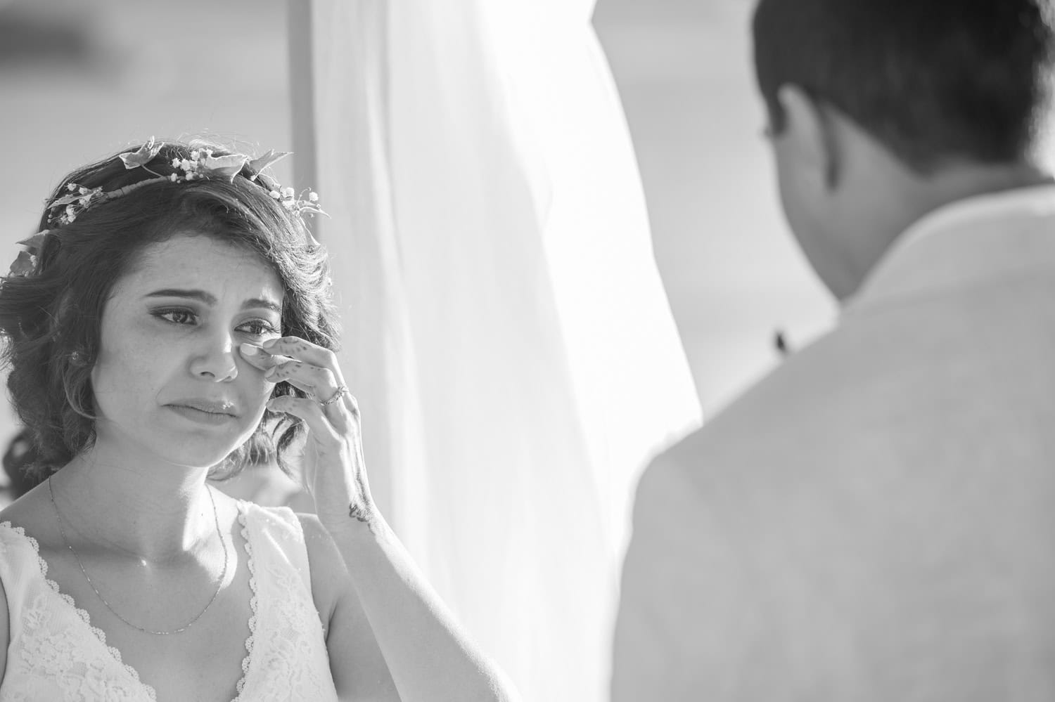 A woman in a wedding dress wipes her eye while facing a man in a light-colored suit. The setting appears emotional.
