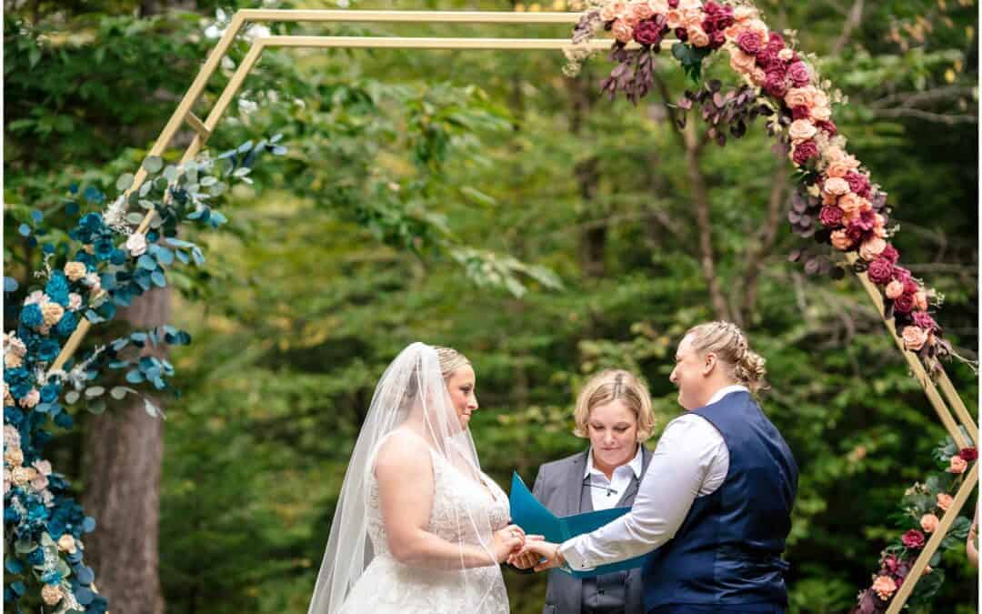 2 people exchange vows in an outdoor wedding ceremony, standing under a hexagonal arch adorned with colorful flowers. An officiant stands between them, holding a document.