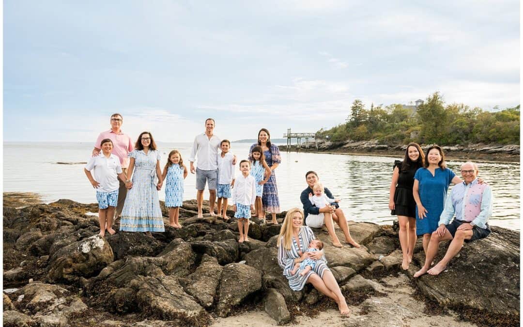 A group of adults and children, stands and sits on a rocky shoreline with the ocean and trees in the background. Some of them are smiling at the camera.