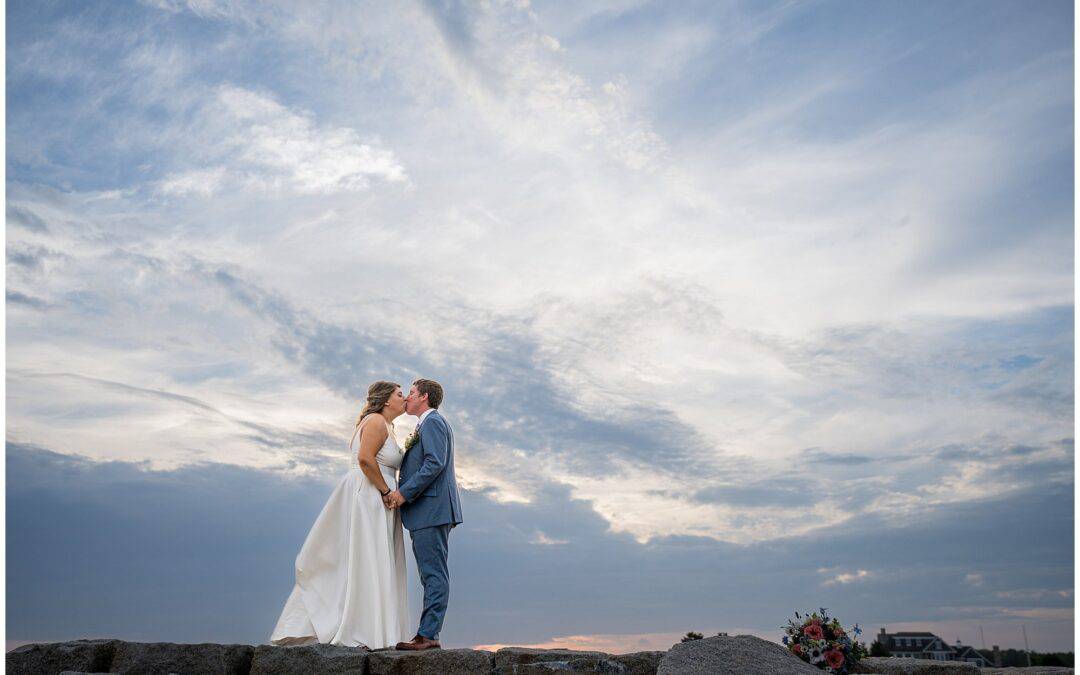 A bride and groom stand on a rocky surface, holding hands and kissing under a dramatic cloudy sky. A bouquet rests on the ground nearby.