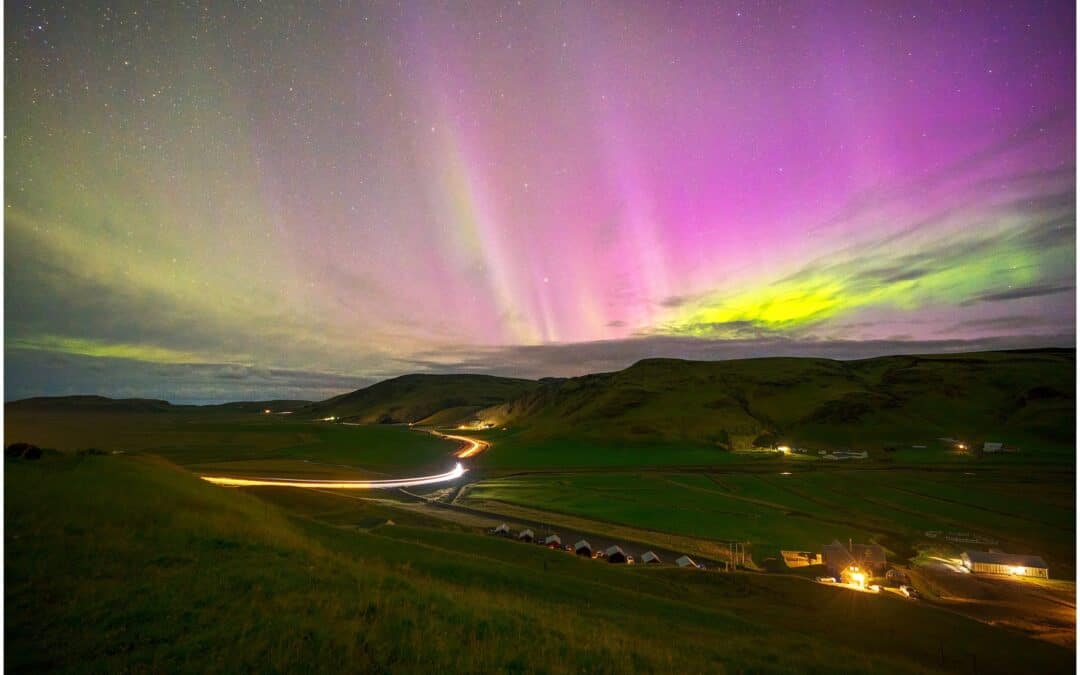 A nighttime landscape features the Northern Lights casting shades of pink and green over rolling hills and a winding road illuminated by car lights.