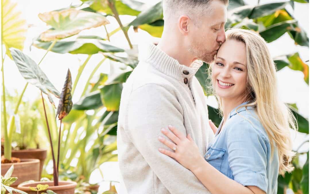 A man kisses a smiling woman on the forehead while embracing her. They are in a setting with abundant green plants and natural light.