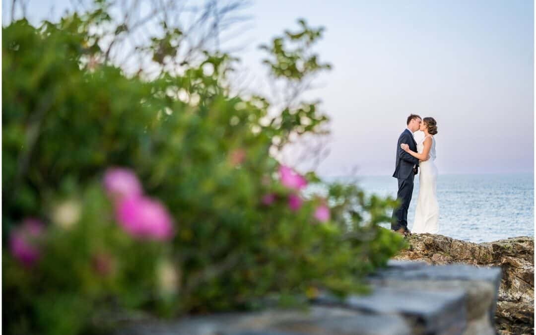 A couple dressed formally stands close together on a rocky shoreline, with an ocean background and flowers in the foreground.