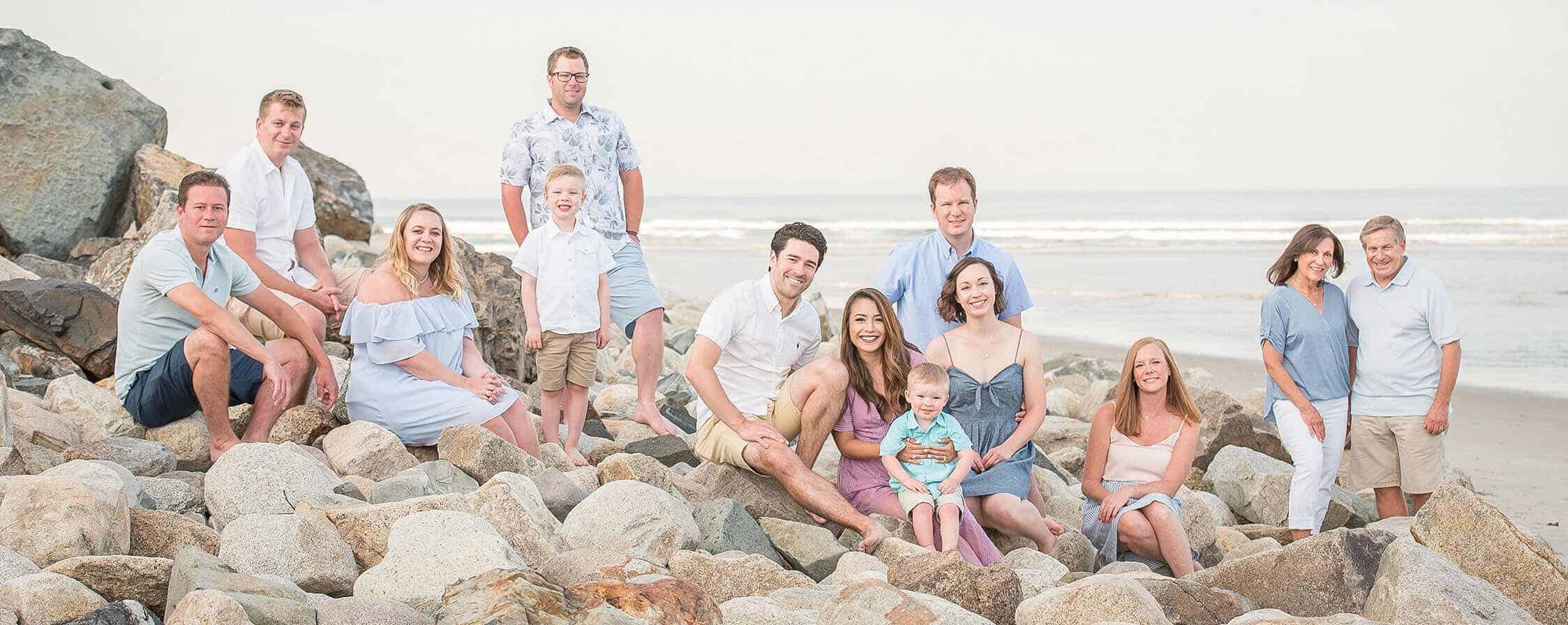 A group of thirteen people, including adults, teens, and young children, pose on rocks at a beach with the ocean in the background on a cloudy day.