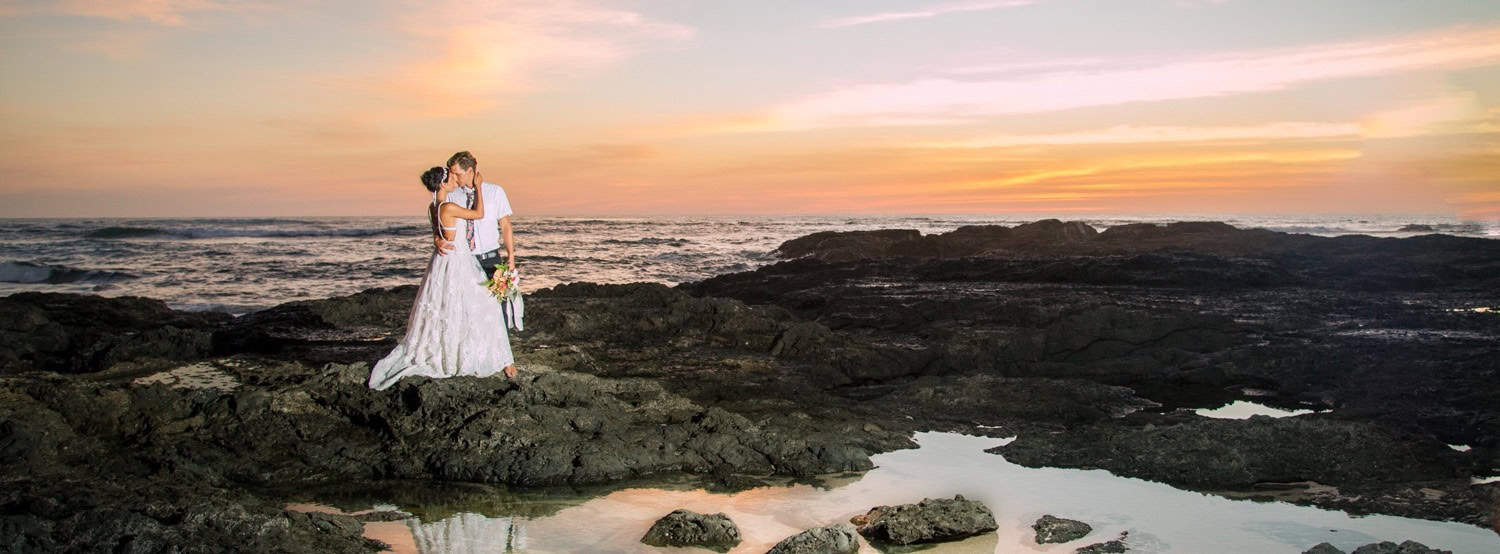 A bride and groom embrace on a rocky beach at sunset, with the ocean and a colorful sky in the background.