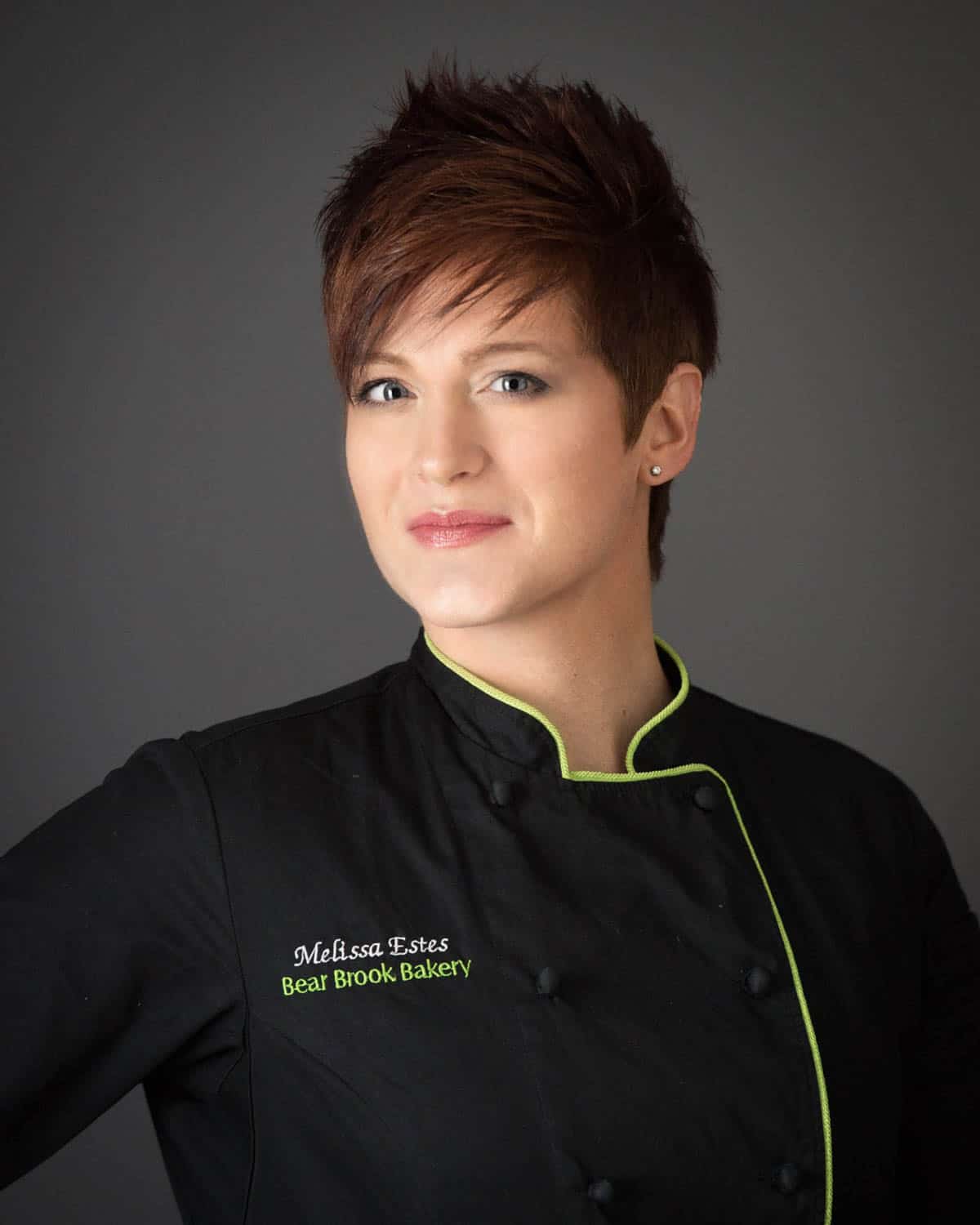 A woman with short hair wearing a black chef's coat with green trim embroidered with Melissa Estes, Bear Brook Bakery stands against a dark background.