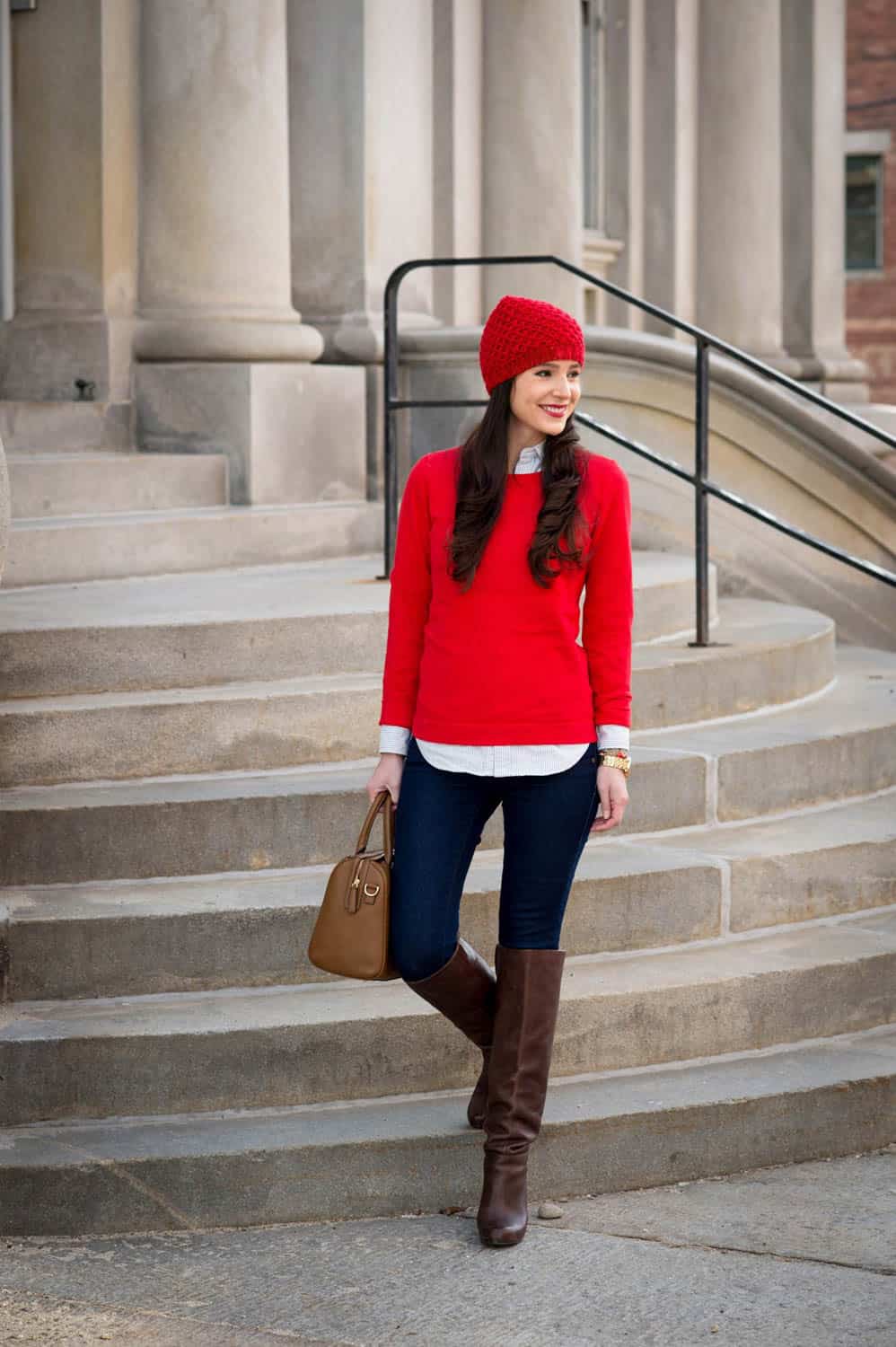 A woman in a red sweater and matching red hat, holding a brown handbag, stands in front of stone steps, wearing dark jeans and brown knee-high boots.