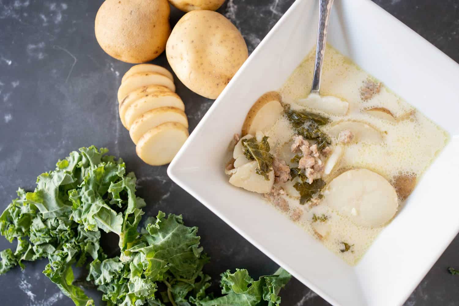 A bowl of creamy soup with potatoes, sausage, and kale sits on a dark counter beside sliced potatoes, whole potatoes, and loose kale leaves.