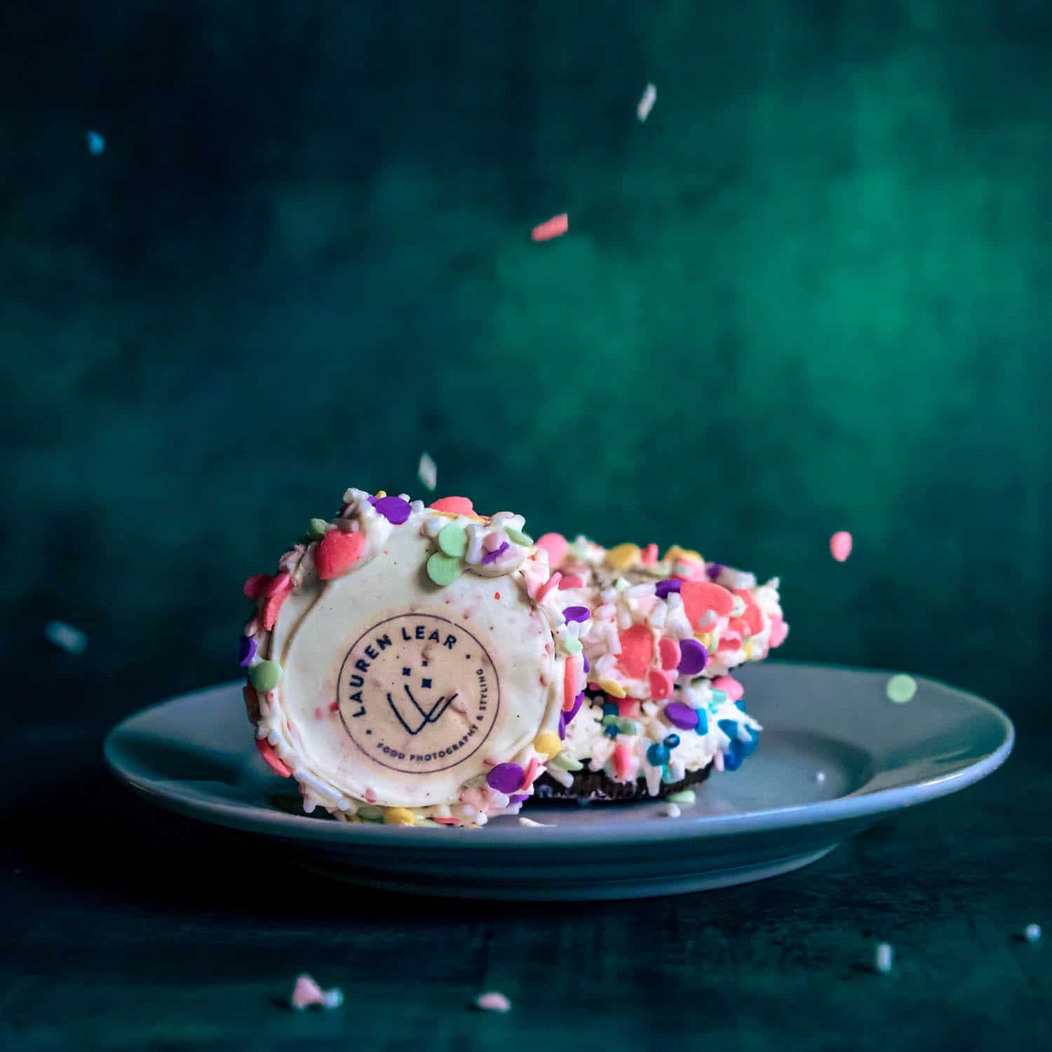 A plate with sprinkle and icing-covered cookies against a green background, one showing a Lauren Lear logo in the center. Sprinkles are falling around the plate.