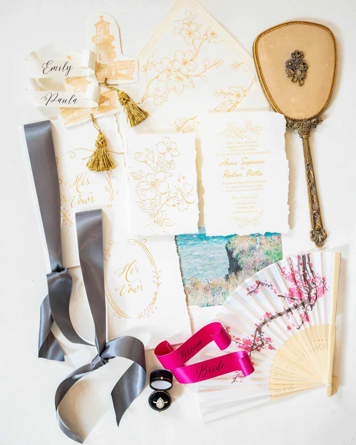 A collection of wedding items including invitations, printed ribbons, rings, decorative fan, ornate hand mirror, tassel, and a botanical print.