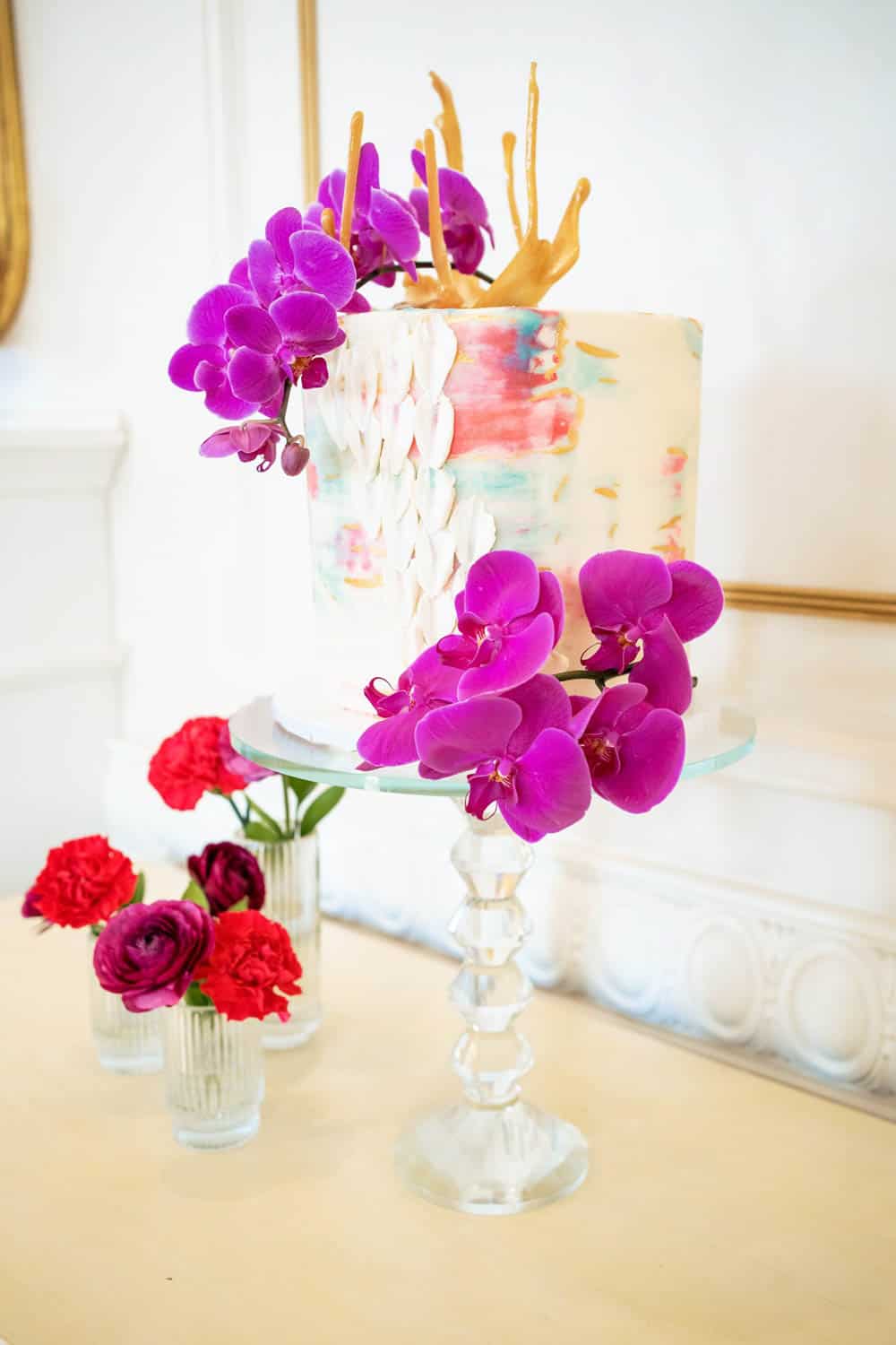 A decorated cake with purple flowers and gold accents sits on a glass stand, accompanied by red flowers in small vases on the table.