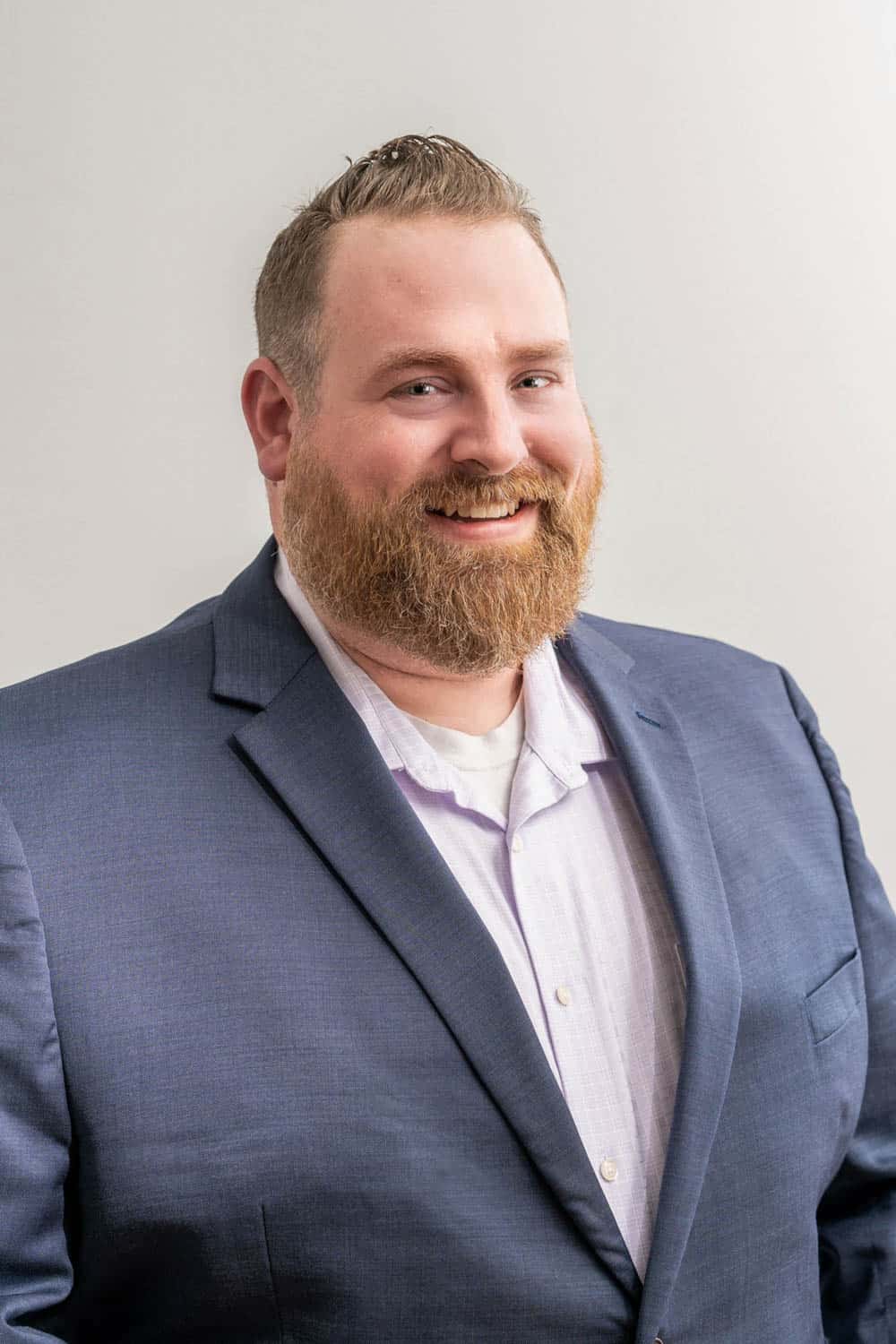 A man with a neatly groomed beard and mustache is wearing a blue suit jacket, a white shirt, and is smiling at the camera against a plain background.