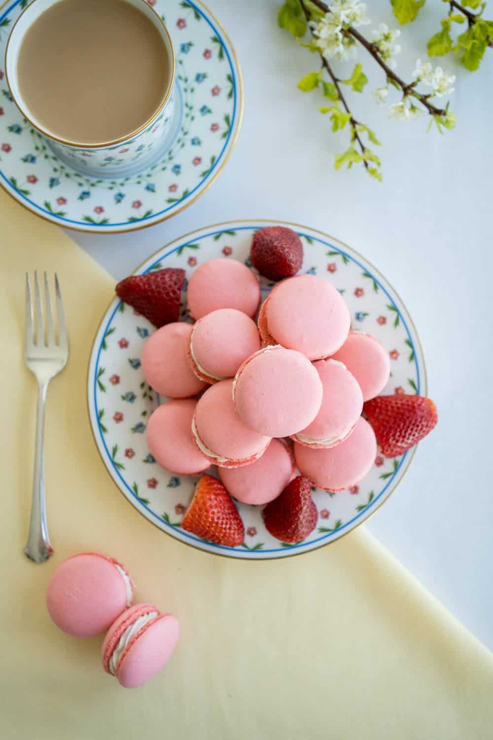 A plate of pink macarons with strawberries, and a cup of coffee in a floral-patterned dish set on a yellow cloth with a fork beside.