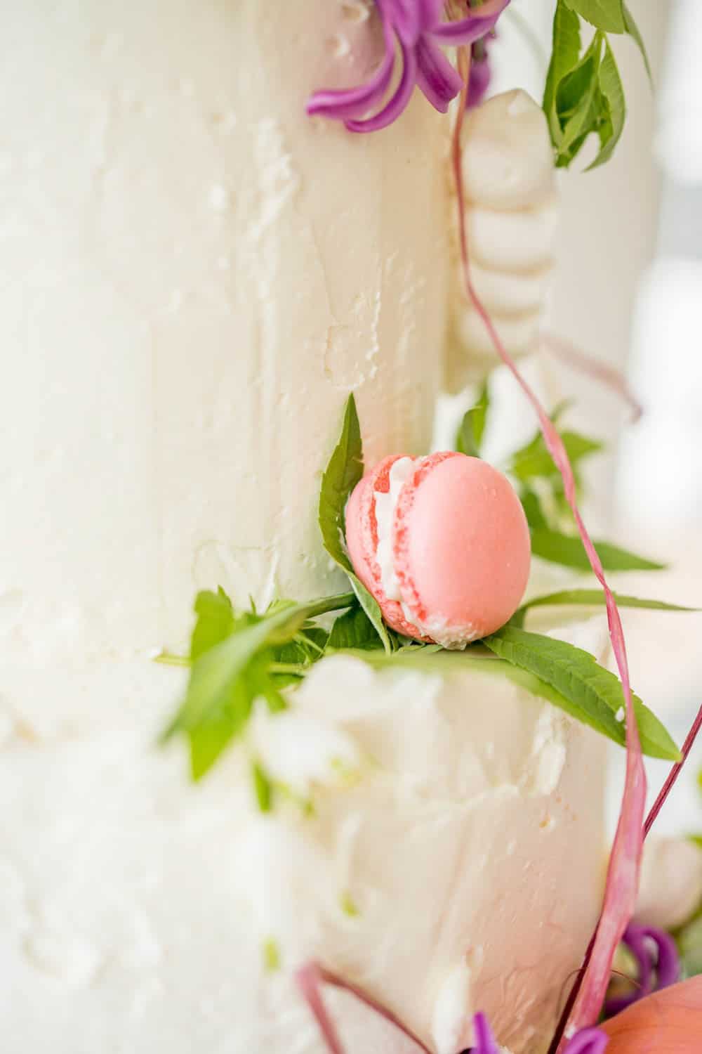 Close-up of a white frosted cake decorated with leaves, pink flowers, and a single pink macaron.