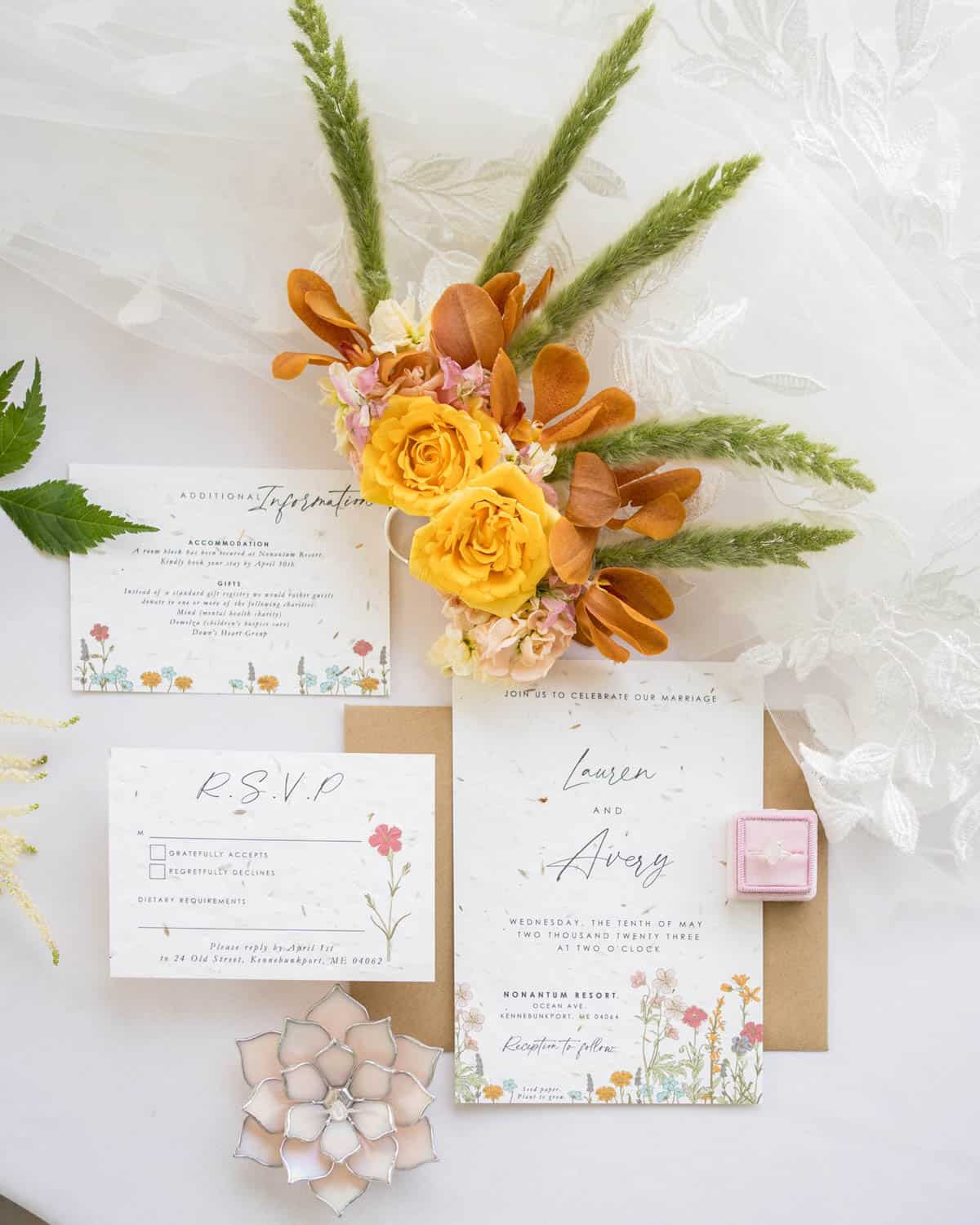 A wedding invitation suite featuring RSVP and details cards, next to a bridal bouquet with yellow and orange flowers, and a ring in an open pink box.