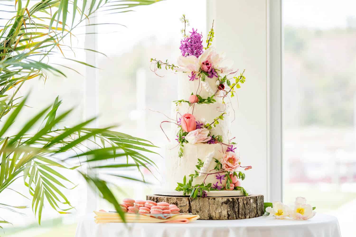 A 3 tier white cake with pink and purple flowers, placed on a wood piece. The cake is set on a round table with a white cloth, green plants, and pink macarons.