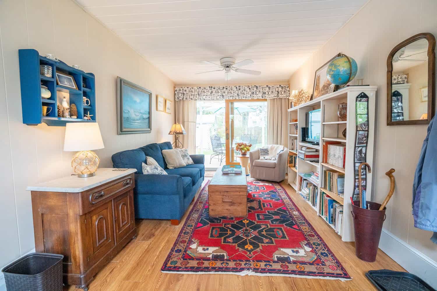 Cozy living room with a blue couch, wood coffee table, white shelves with books and a TV, ornate rug, and sliding glass door leading outside.