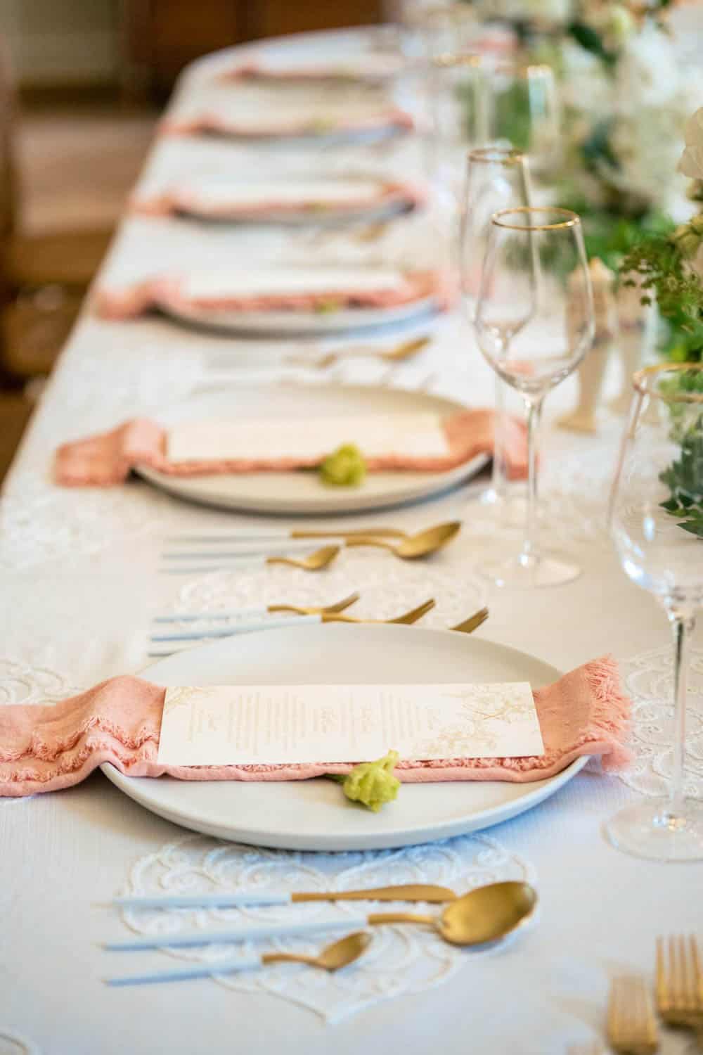 Elegant table setting with white plates, pink napkins, menus, gold cutlery, and wine glasses on a white lace tablecloth.