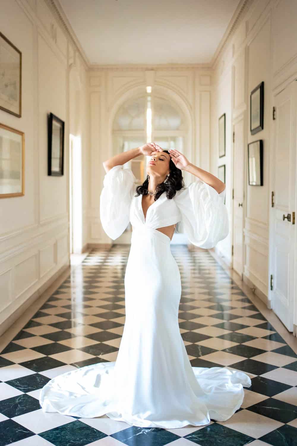 A woman wearing a formal white gown with puffed sleeves stands in a hallway with checkered flooring, striking a dramatic pose.