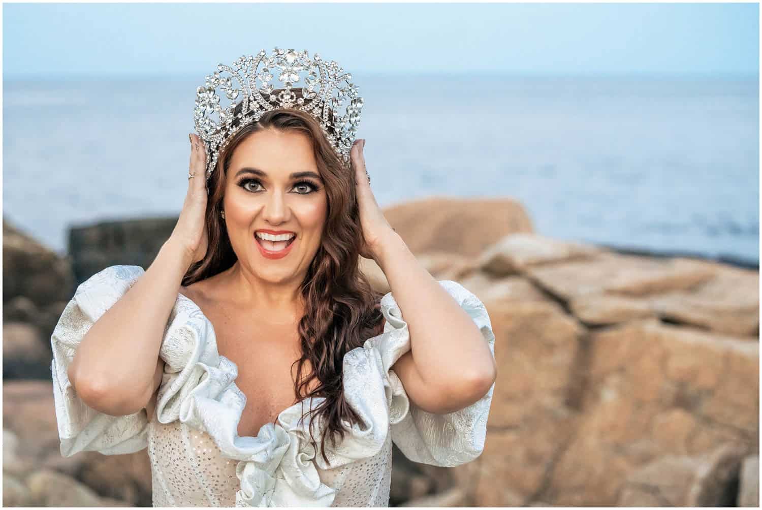 A woman smiles broadly while holding a jeweled crown on her head. She is wearing a white dress with ruffled sleeves, standing in front of a rocky shoreline.