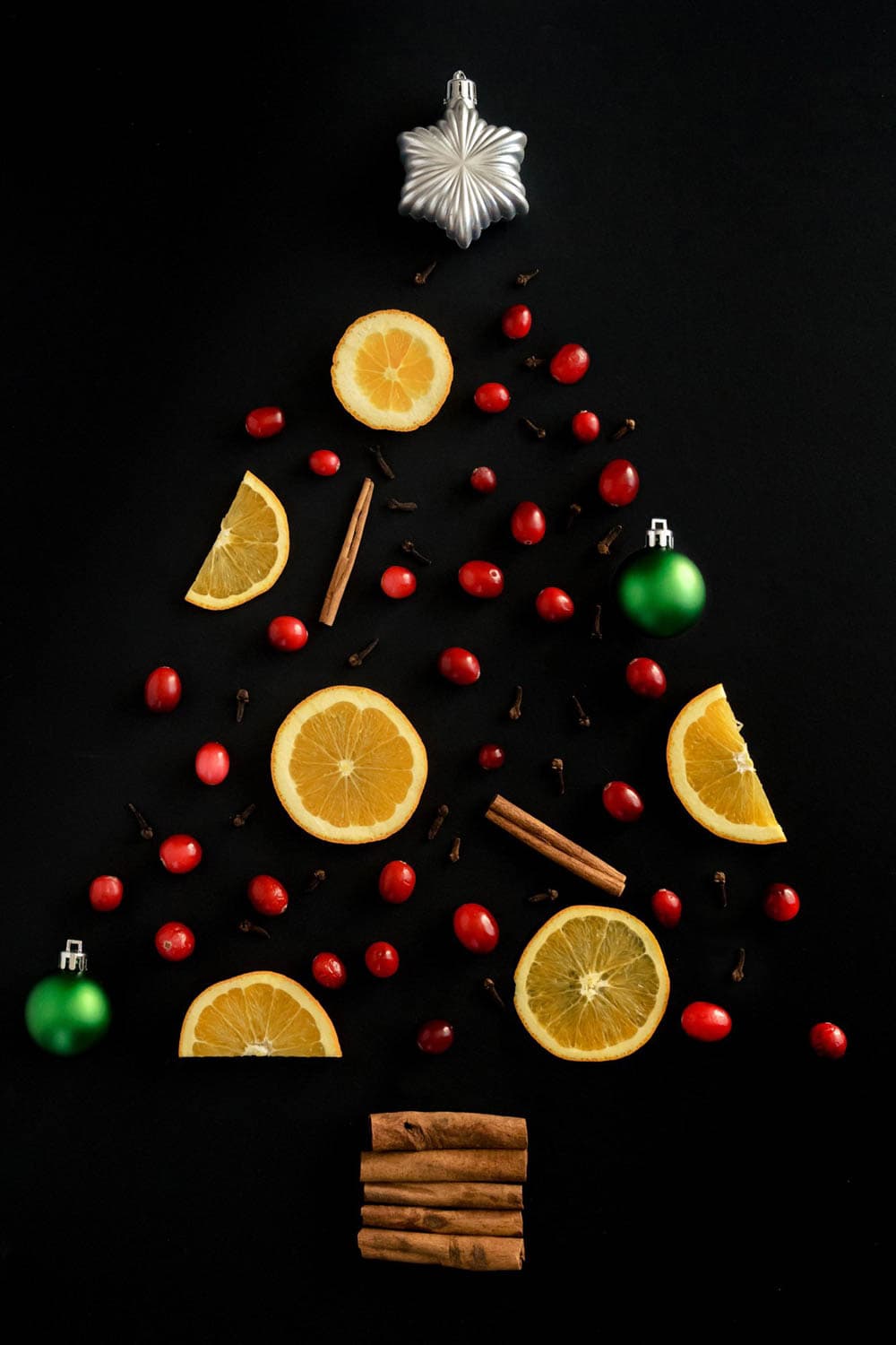 A Christmas tree design made with orange slices, cranberries, cinnamon sticks, cloves, and green ornaments on a black background.