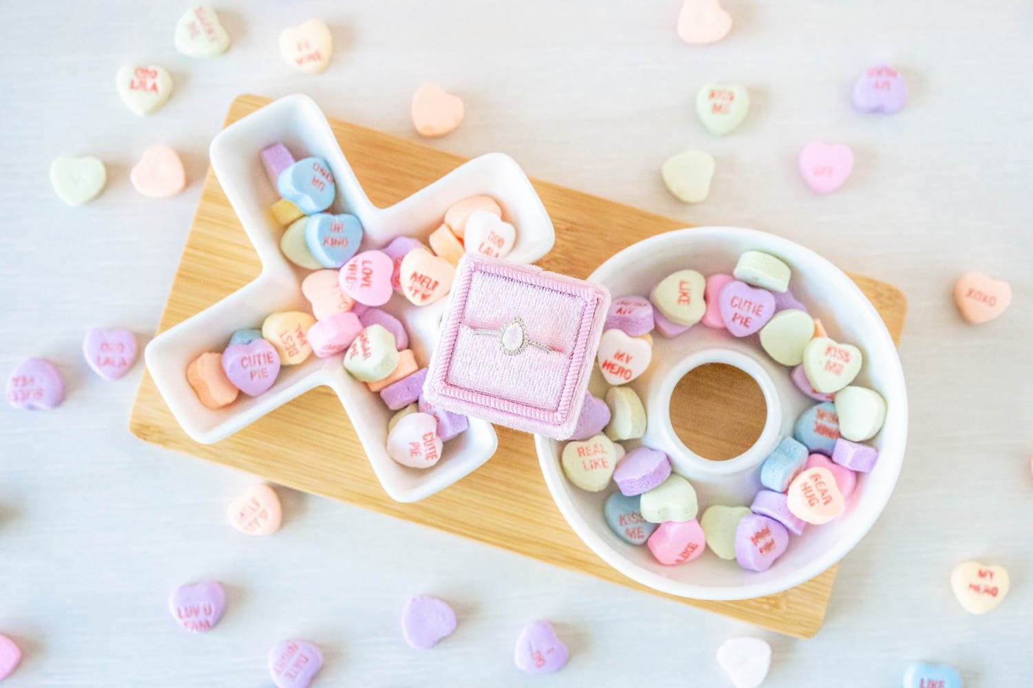 A wood tray holds heart-shaped candies in pastel colors with messages. A ring box with a diamond ring is placed among the candies, signifying a proposal.