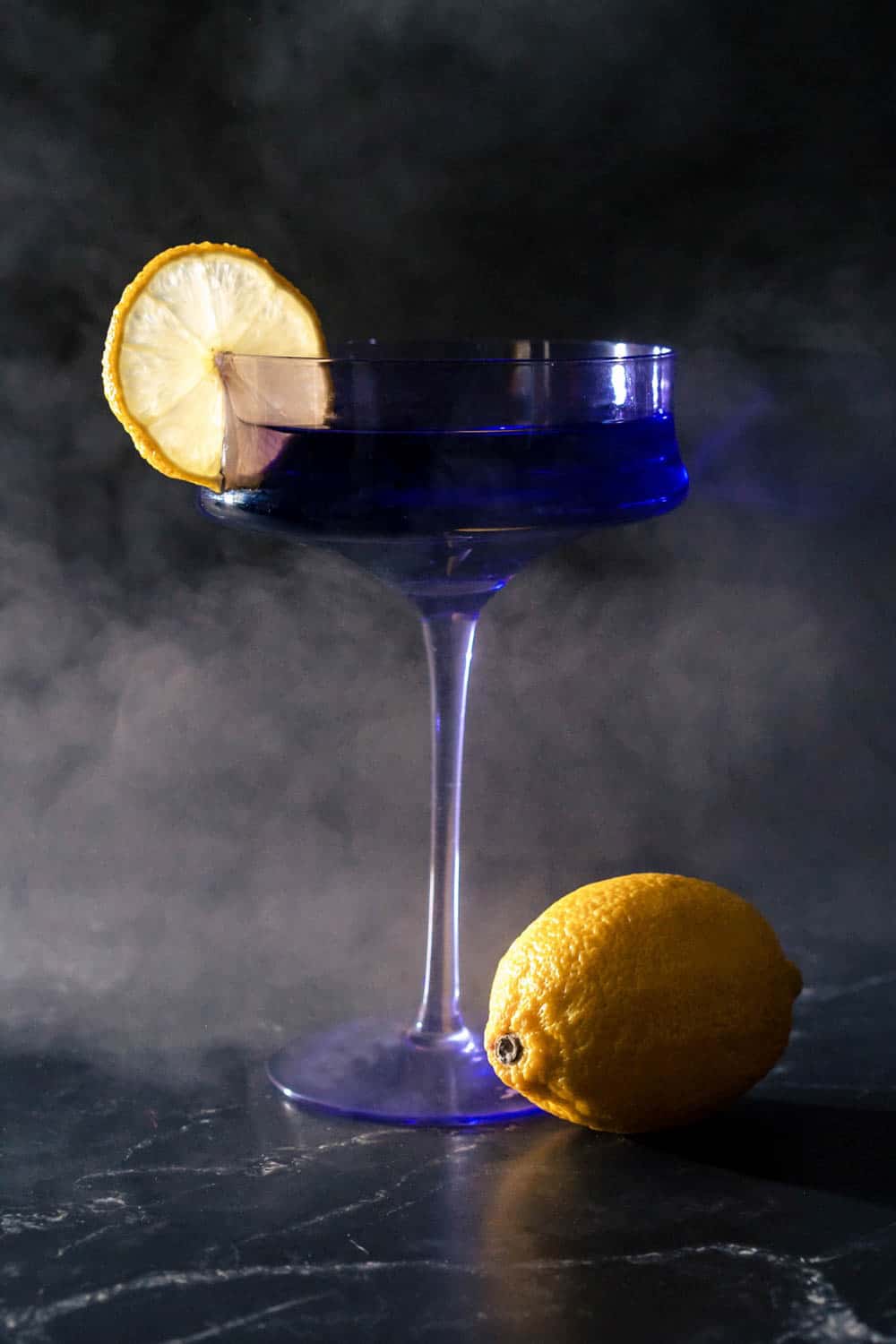 A blue cocktail in a glass garnished with a lemon slice. A whole lemon is placed next to the glass on a dark smoky background.