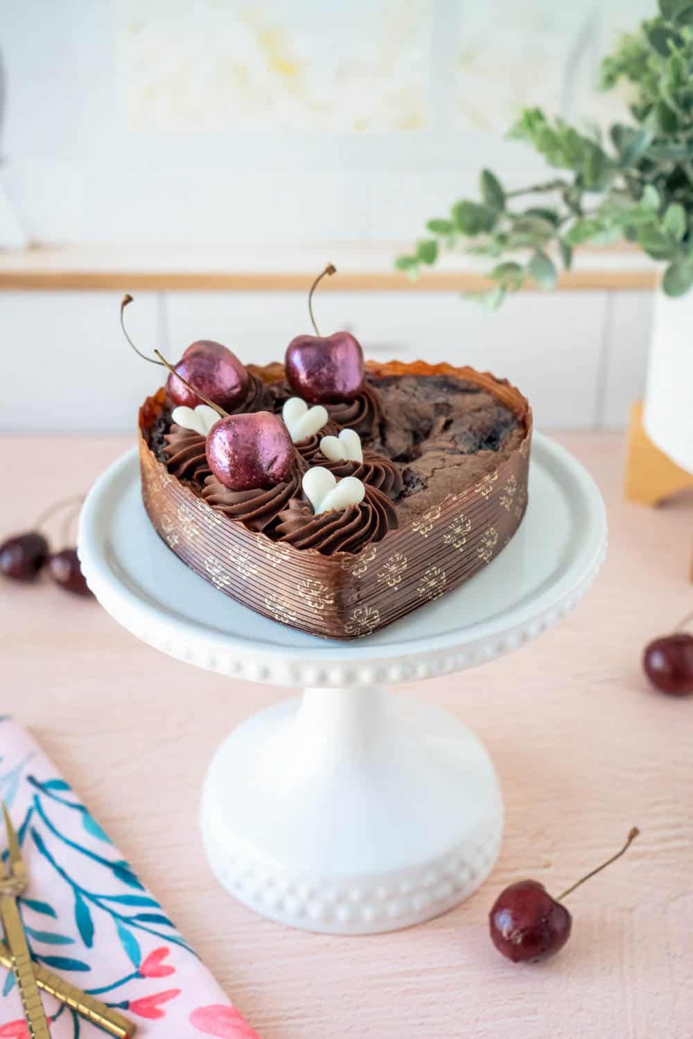 Heart-shaped chocolate cake topped with cherries and white chocolate garnishes, on a white pedestal cake stand. Additional cherries are on the table.