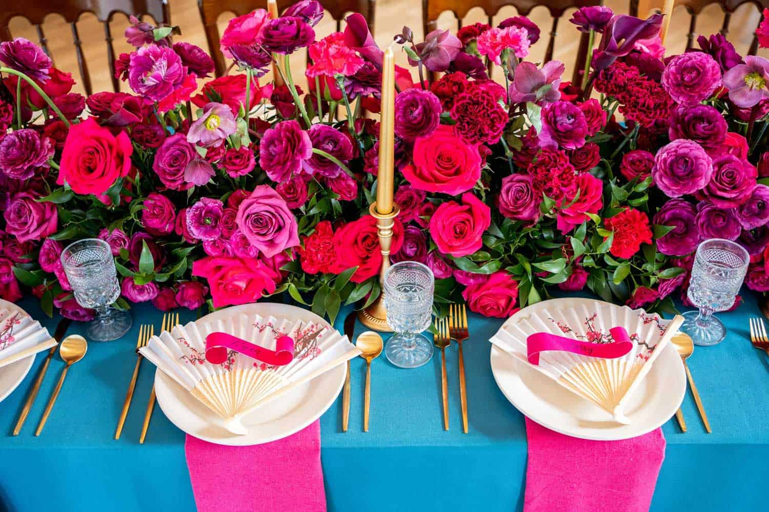 A vibrant floral arrangement featuring red and pink flowers decorates a dinner table set with white plates, gold cutlery, crystal glasses, and teal tablecloths with pink napkins.