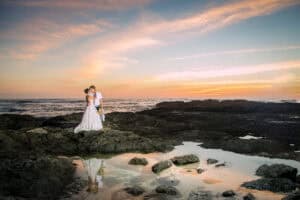 A bride and groom stand on rocky terrain near the ocean at sunset, with the sky displaying shades of orange and pink.