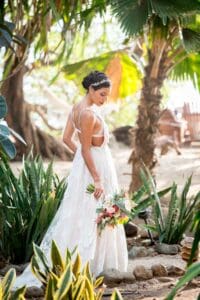 Bride in a white gown holding a bouquet of flowers, surrounded by tropical plants.