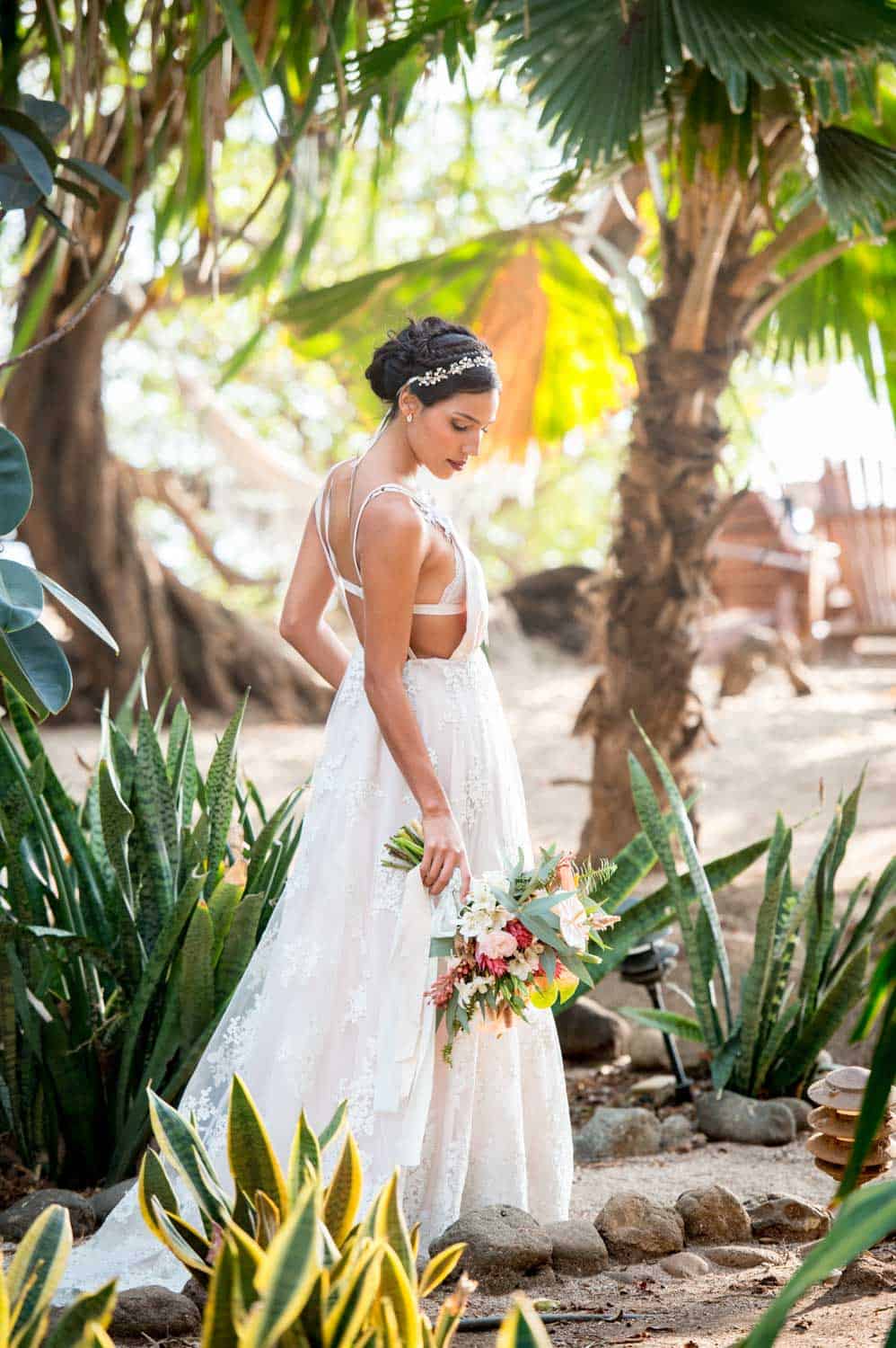 Bride in a white gown holding a bouquet of flowers, surrounded by tropical plants.