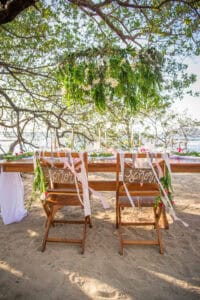A decorated outdoor dining table with two wooden chairs labeled Señor and Señora adorned with ribbons and greenery under a tree near the beach.