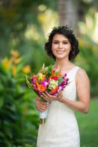 A woman in a sleeveless white dress holds a vibrant bouquet of flowers, standing outdoors with green foliage in the background.