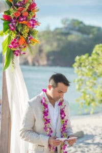 A man in a light-colored suit adorned with a flower lei reads from a piece of paper beneath a tropical floral arrangement by the beach.