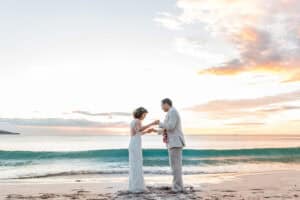 A couple in wedding attire stands on a beach at sunset, exchanging leis while facing each other with the ocean and sky in the background.
