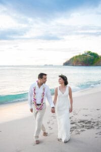 A couple dressed in white wedding attire, with the groom wearing a floral lei, walk hand-in-hand along a beach.