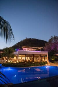 A modern building with string lights on the rooftop stands next to a blue-lit swimming pool during twilight, surrounded by palm trees and hilly terrain.