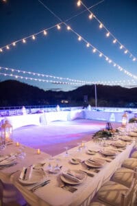 Outdoor dinner setup with long tables covered in white tablecloths, plates, utensils, glasses, centerpieces, and lanterns under string lights at sunset.