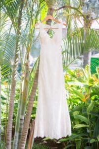 A sleeveless white lace wedding dress hangs from a wooden hanger amidst green tropical plants.