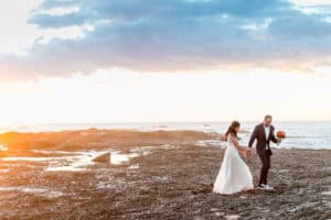 A bride and groom walk on a rocky shore at sunset, the groom holding a bouquet of colorful flowers.