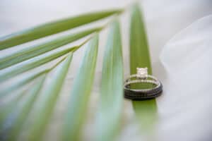 Close-up of two rings resting on green palm leaves. The top ring has a diamond, and they are placed on a light, sheer fabric background.