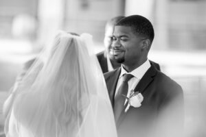 A groom smiles at his bride during their wedding ceremony. The couple is facing each other, with the bride wearing a veil and the groom in a suit.