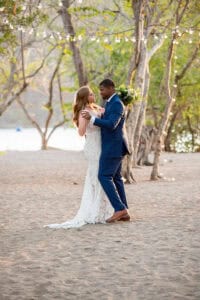 A couple, dressed in wedding attire, is embracing under string lights in a wooded outdoor setting with a lake in the background.