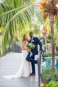 A bride and groom pose together on a wood bridge surrounded by palm trees, with the groom leaning on the railing and the bride holding a bouquet of white flowers.