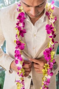 A man wearing a beige suit jacket and a white shirt adjusts the buttons. He has a lei of pink, purple, and green flowers draped around his neck.