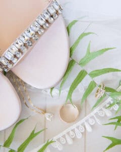 Close-up of elegant shoes with jeweled embellishments, a teardrop pendant necklace, a gold wedding band, and an engagement ring, on a leaf-patterned fabric.