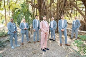 A group of seven men dressed in suits, with the groom in a pink suit standing in the center, pose outdoors under large, intertwined tree branches.